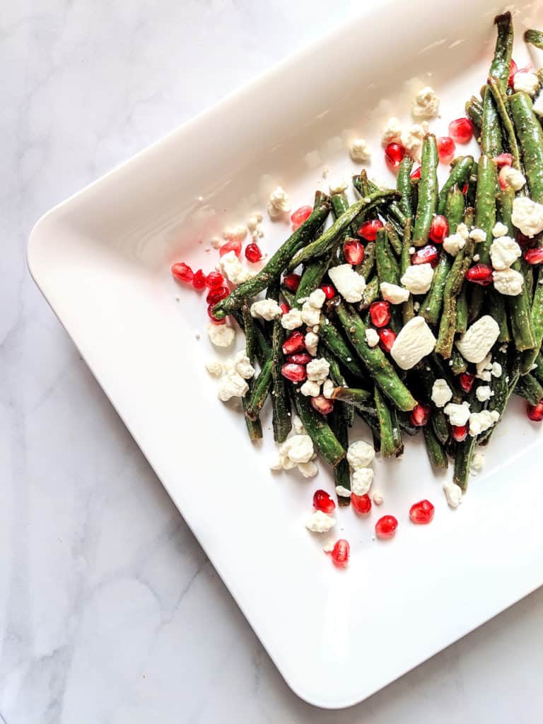 roasted green beans with crumbled goat cheese and pomegranate seeds on a white rectangular plate and light grey background